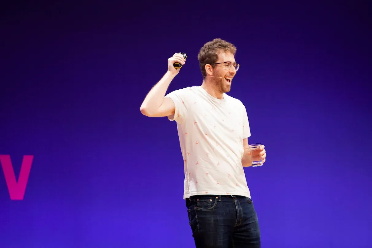 Me laughing and speaking with a conference mic on my face, wearing a white t-shirt with small pink flamingos, holding a cup of water